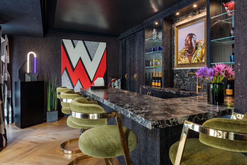 Modern home bar with green velvet stools, marble countertop, contemporary artwork, neon lamp, and a portrait on the wall.