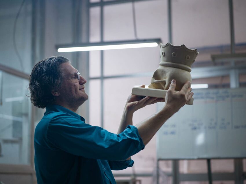 Man in blue shirt holding up a ceramic piece shaped like a crown in a workshop setting.