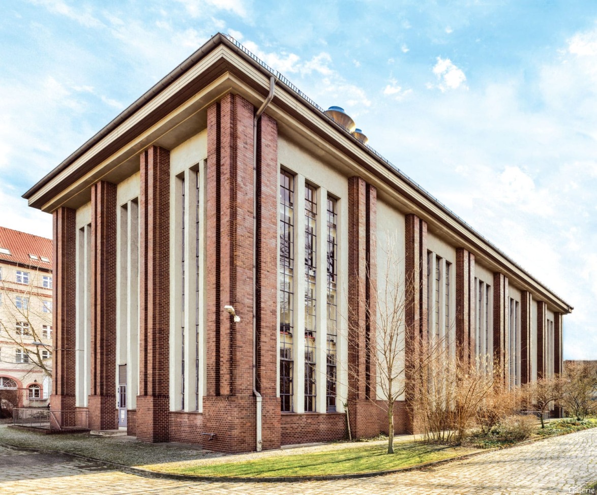 Red-brick and beige stone building with tall vertical windows surrounded by pavement and trees under a partly cloudy sky.