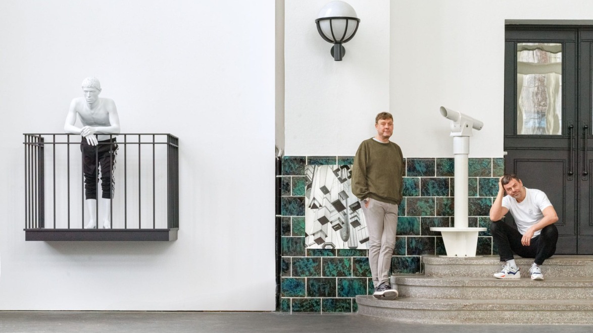 Two men pose near a wall sculpture of a man on a balcony, with steps, a telescope, and tiled wall art in the background.