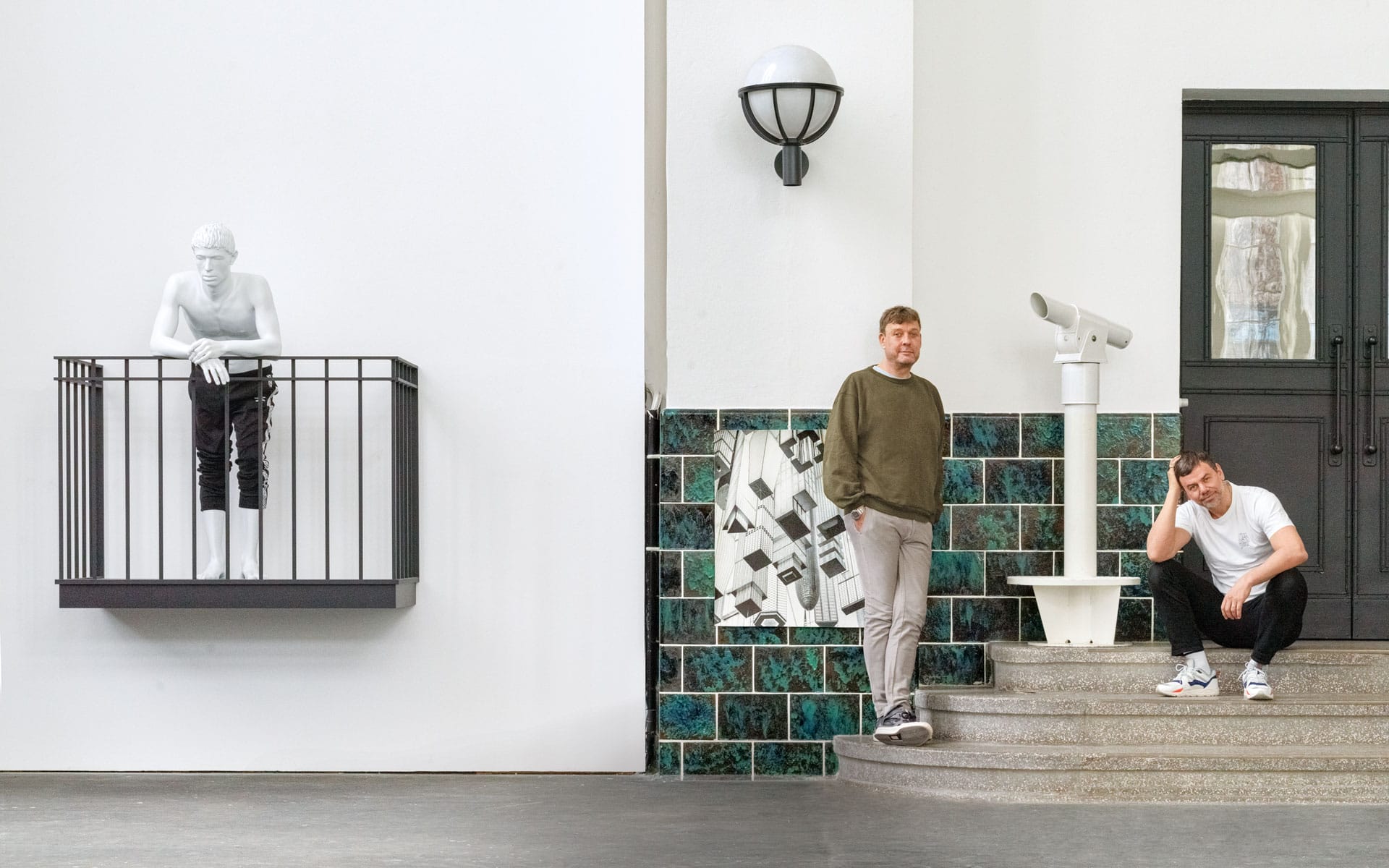 Two men pose near a wall sculpture of a man on a balcony, with steps, a telescope, and tiled wall art in the background.