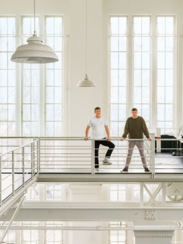 Two people standing on an indoor balcony with large windows in the background, industrial-style lighting overhead.