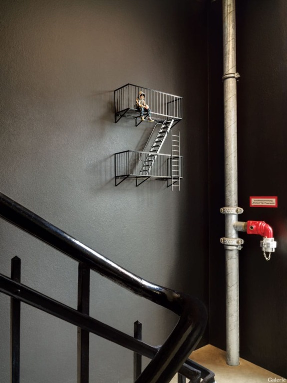 Art installation of a miniature fire escape attached to a black wall with a small figure sitting on it.