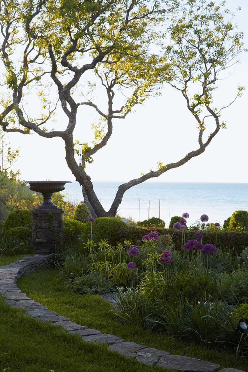 Garden path with blossoming trees and purple flowers, overlooking a calm sea under a clear sky.