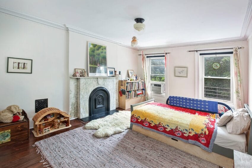 Cozy children's bedroom with a bed, colorful quilt, toy house, fireplace, bookshelf, and large windows letting in natural light.