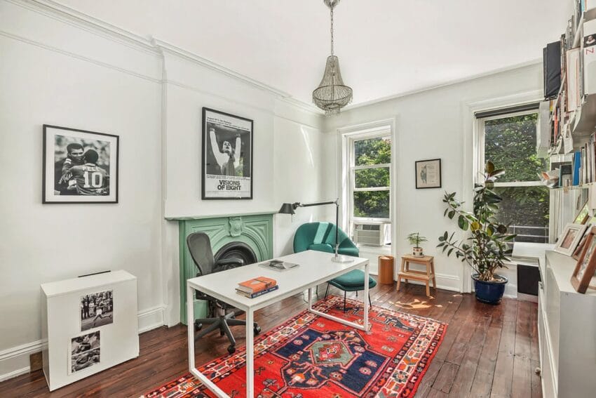 Home office with white walls, desk, chairs, bookshelf, and bright red rug with natural light from windows.