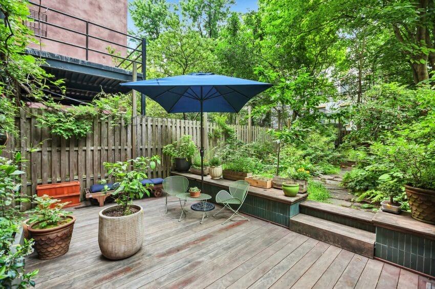 Backyard patio with plants, a blue umbrella, wooden decking, and green chairs surrounded by lush trees and greenery.