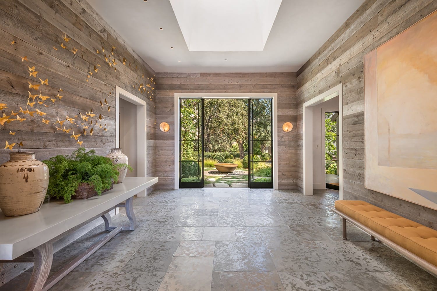 Modern hallway with rustic wooden walls, decorative vases, butterfly wall art, and a view of a lush garden through glass doors.