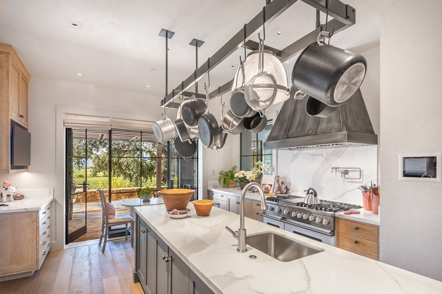 Modern kitchen with marble countertops, hanging pots, stainless steel appliances, and wooden accents, overlooking outdoor patio.