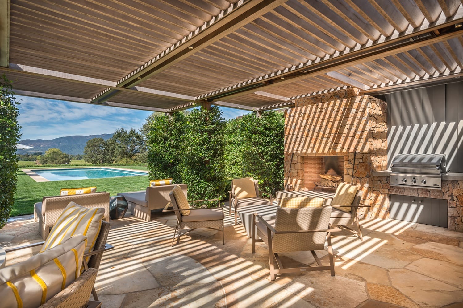 Outdoor patio with wicker furniture and stone fireplace overlooking a pool and mountains under a pergola with sunlight streaming through.
