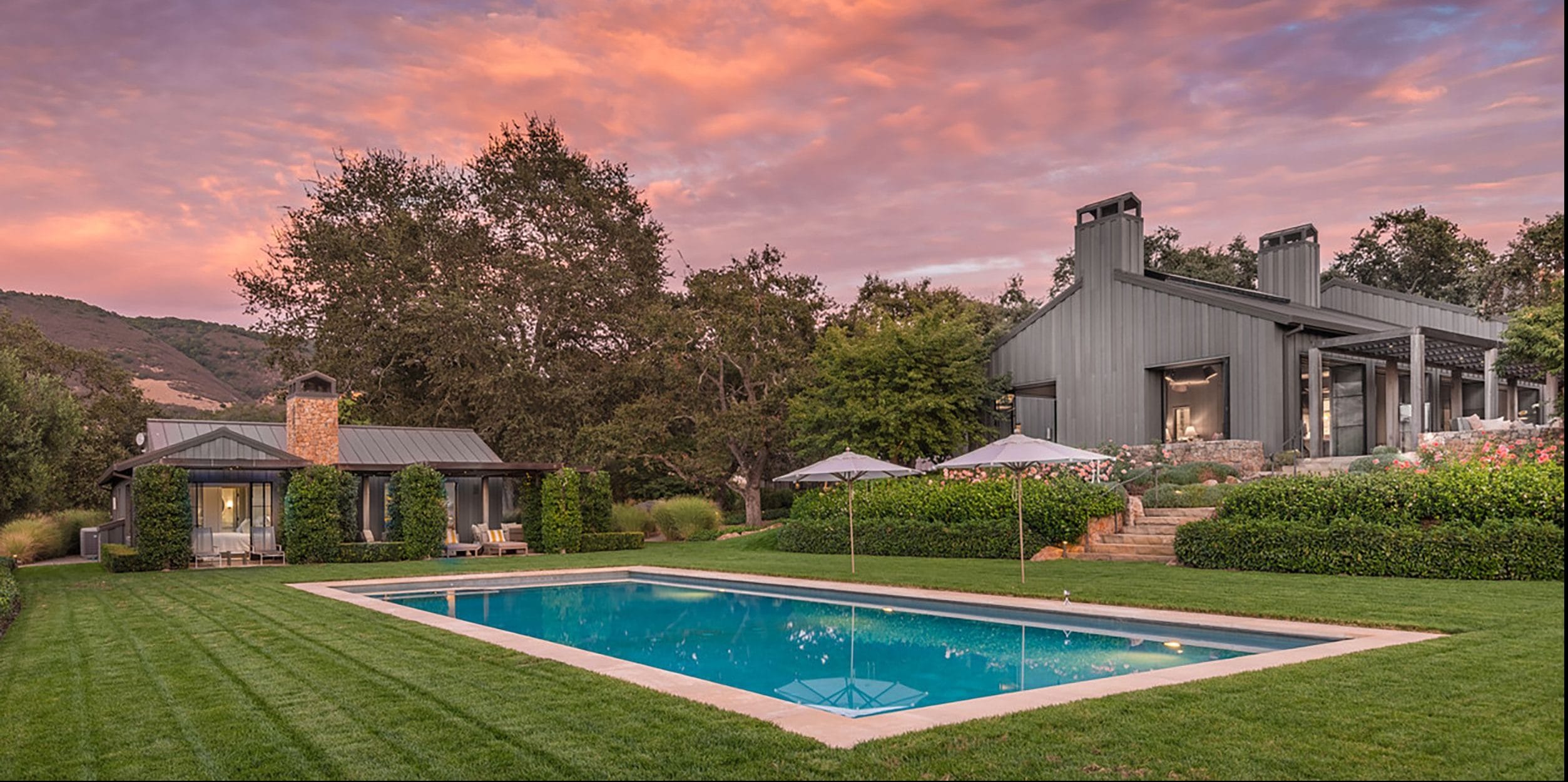 Modern home with large windows, green lawn, and a swimming pool under a vibrant pink and orange sunset sky.