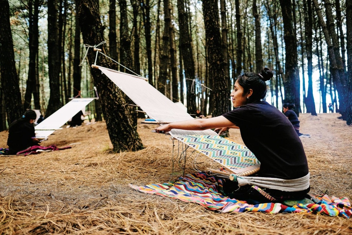 Person weaving on a traditional loom in a forest, sitting on a colorful blanket surrounded by pine trees.