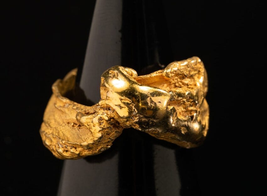 Close-up of a textured gold nugget ring displayed on a black stand against a dark background.