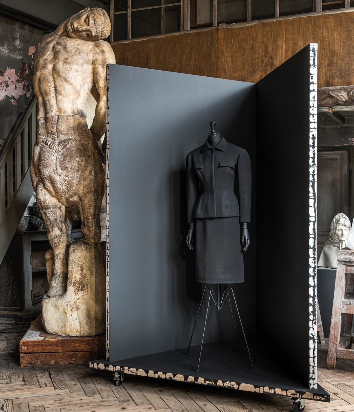 Mannequin in a minimalist black suit displayed in an open box surrounded by statues and art studio elements.