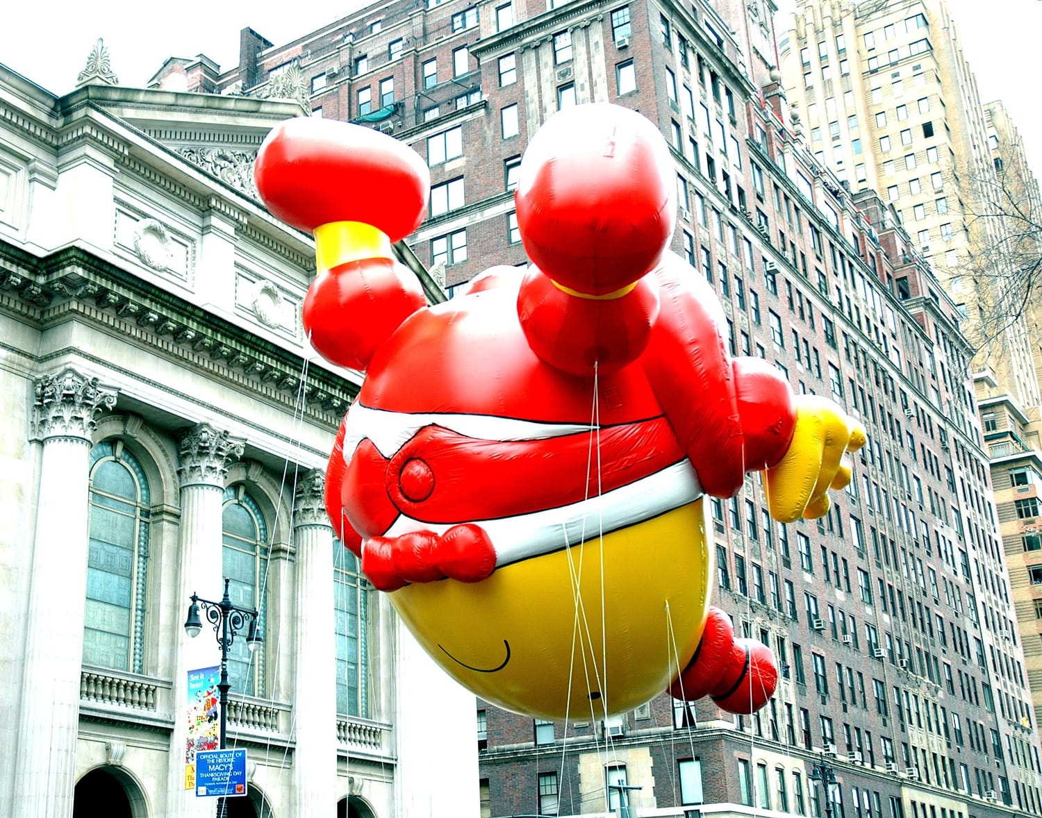 Large red balloon character floats in a city parade, surrounded by tall buildings and spectators.