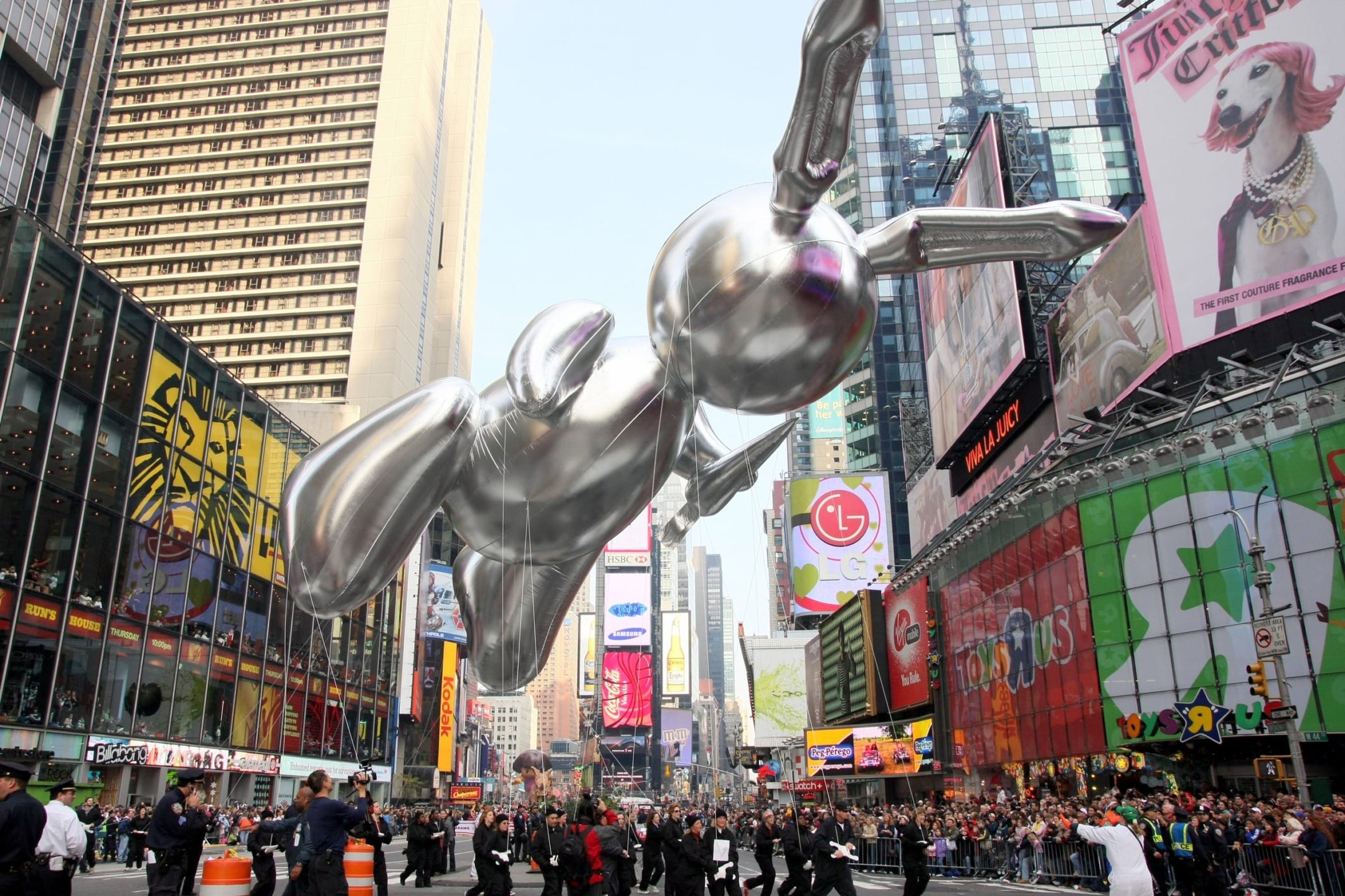 Large silver balloon shaped like a rabbit floats above a crowded city street with tall buildings and colorful billboards.