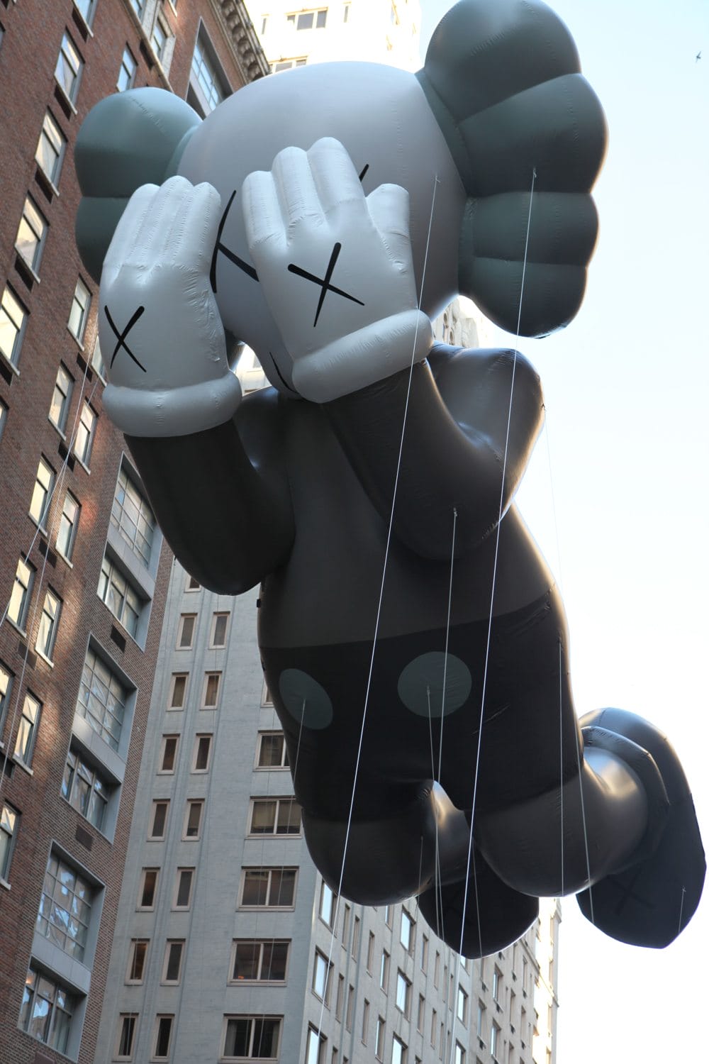 Giant balloon figure with crossed-out eyes and gloved hands covering face, floating between city buildings.
