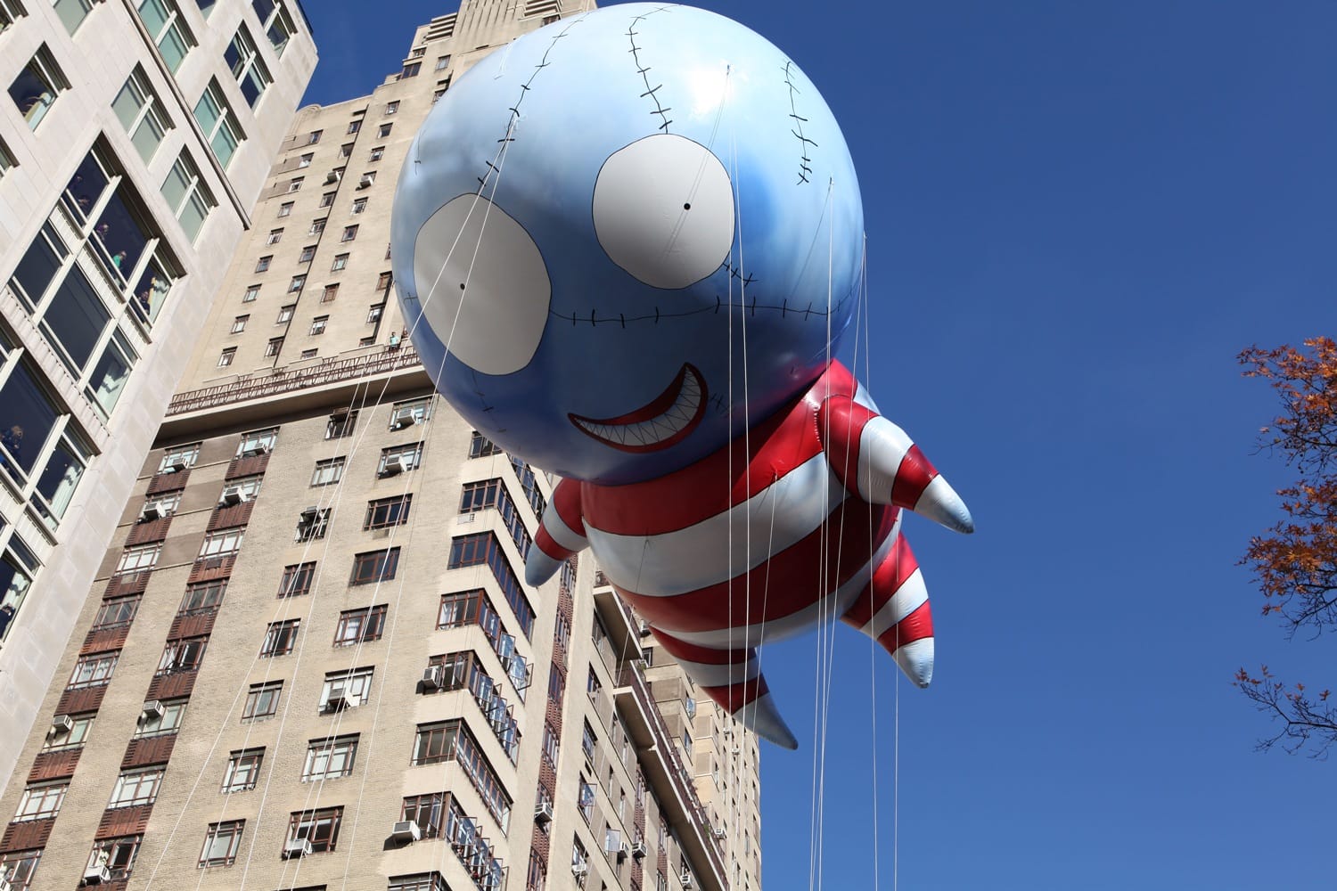 Giant cartoon balloon of blue character with striped outfit floating in New York City during a parade near tall buildings.