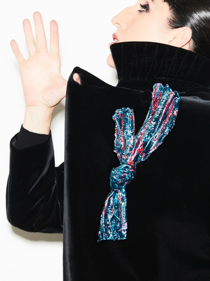 Person in a black velvet jacket with a colorful scarf tied at the back, raising one hand against a white background.