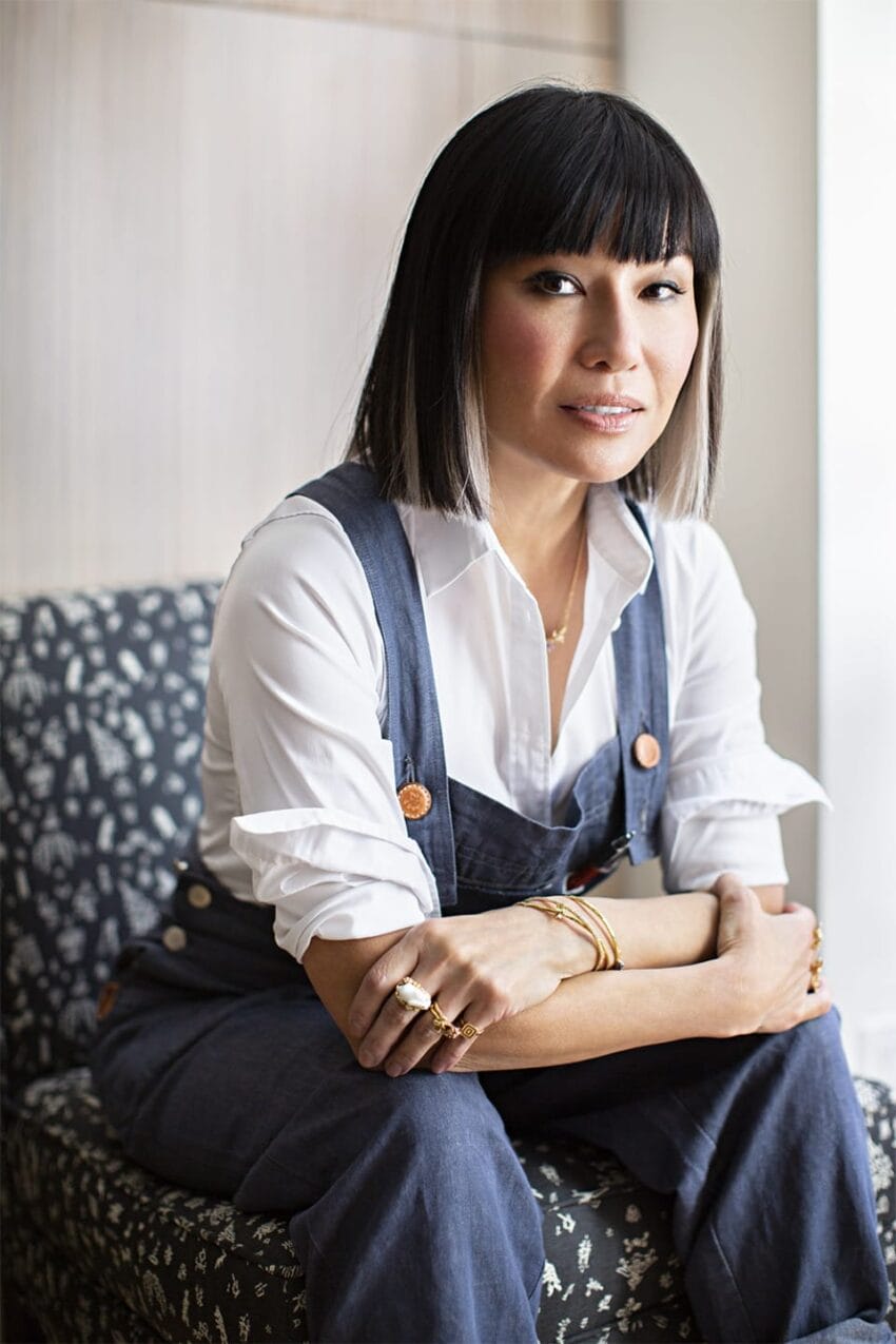 Person sitting on a patterned chair wearing a white shirt and denim overalls, with arms crossed and a neutral expression.