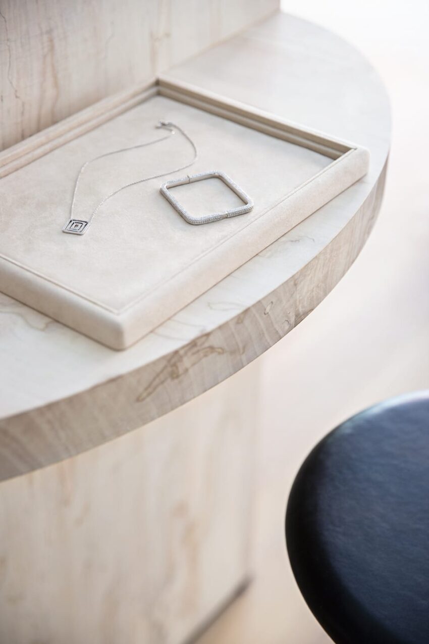 Silver necklace with square pendant and chain on a beige jewelry tray, placed on a wooden table near a black stool.