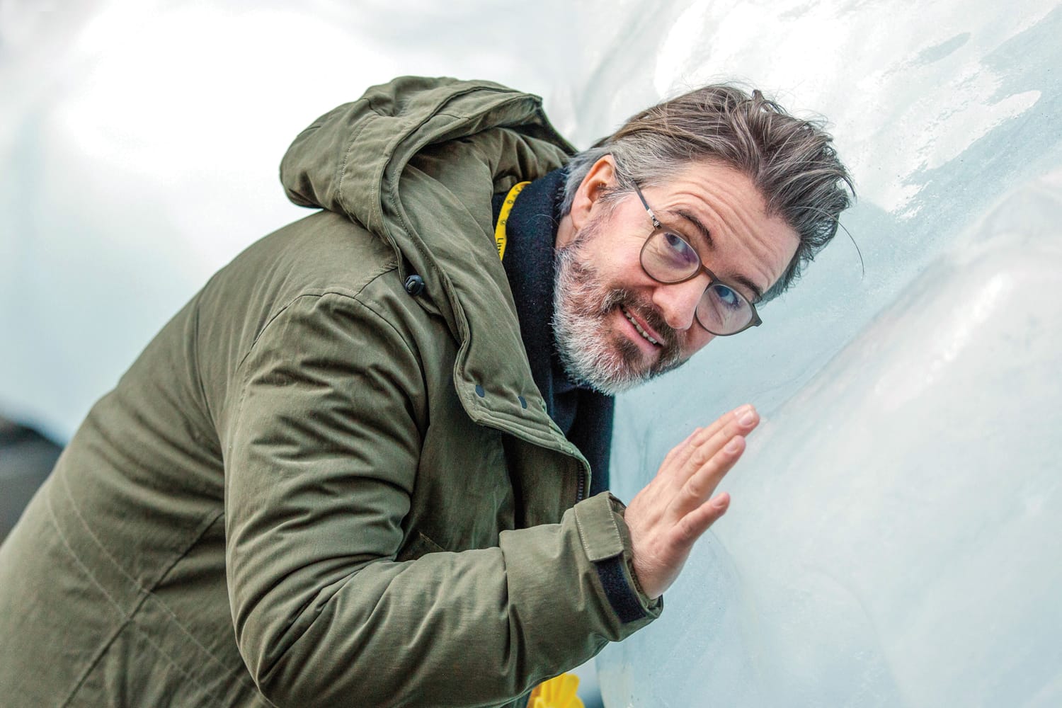 Person wearing glasses and a green coat touching a large block of ice, appearing thoughtful and engaged with the environment.