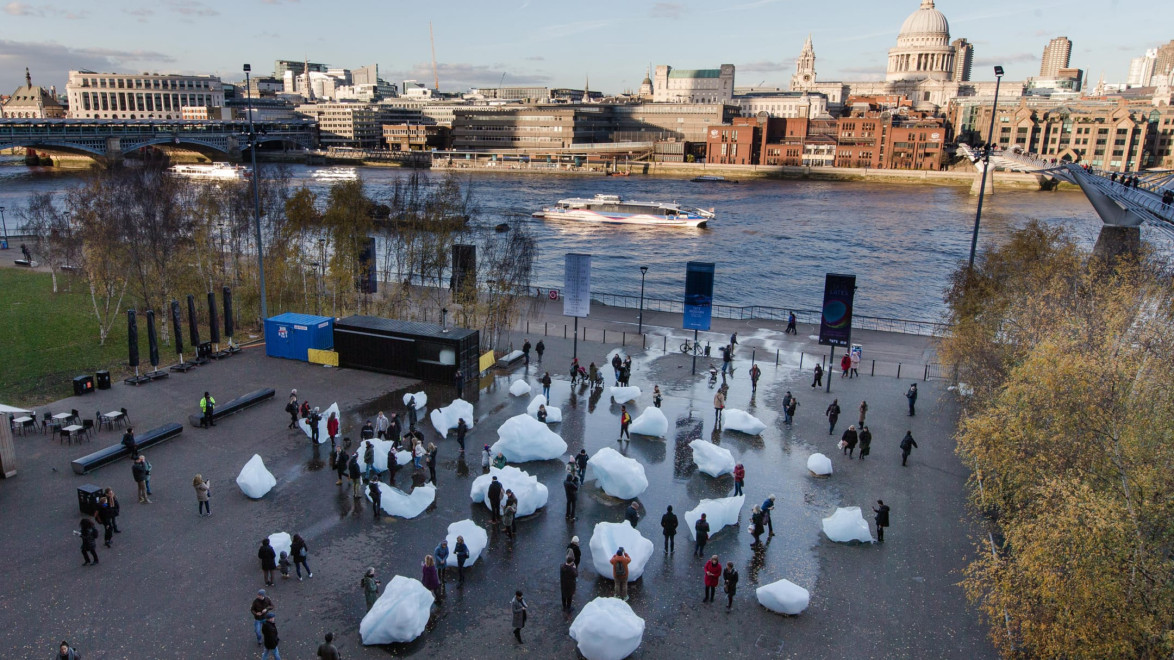 Public art installation with people interacting around large ice blocks near a river, city skyline in the background.