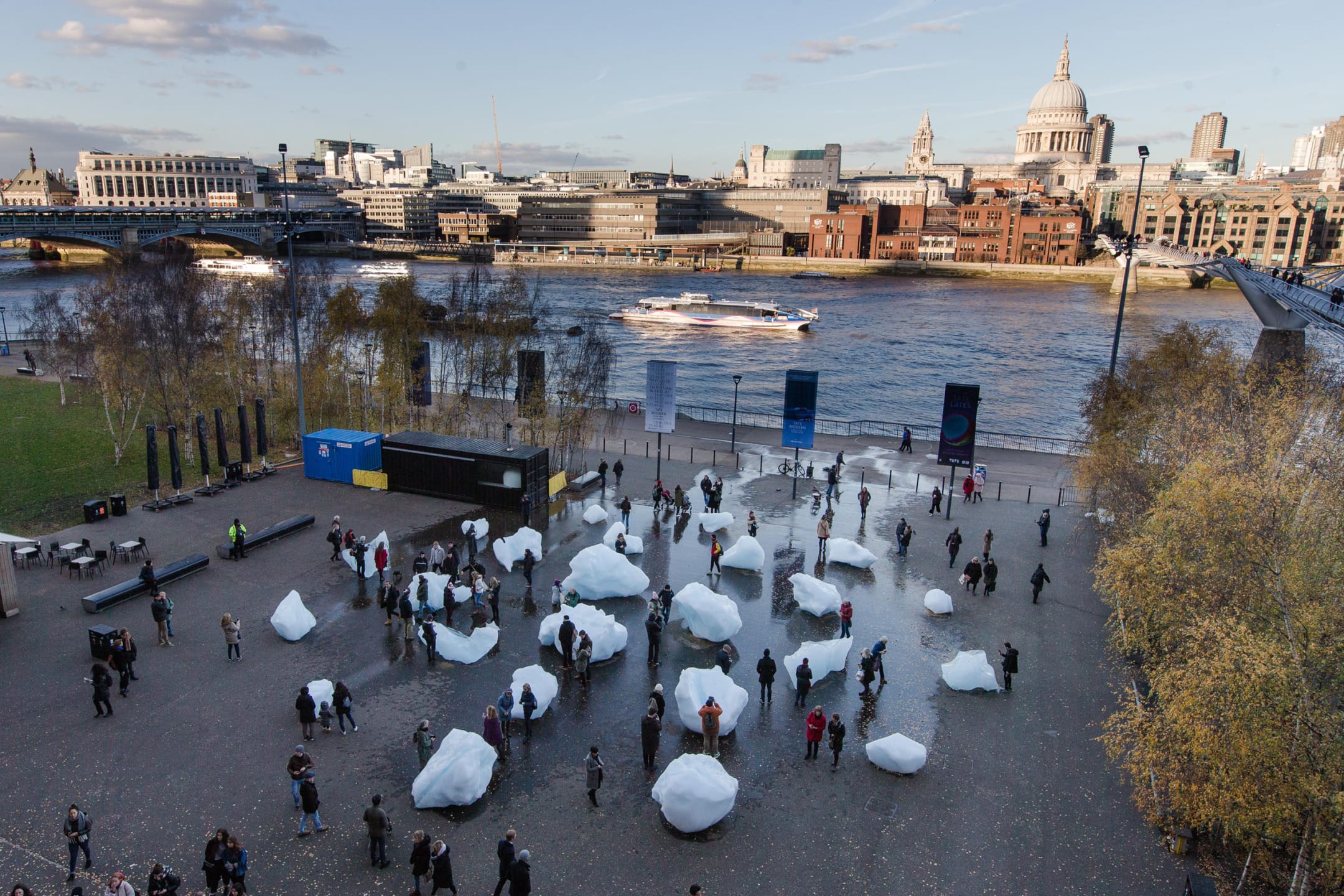 Public art installation with people interacting around large ice blocks near a river, city skyline in the background.