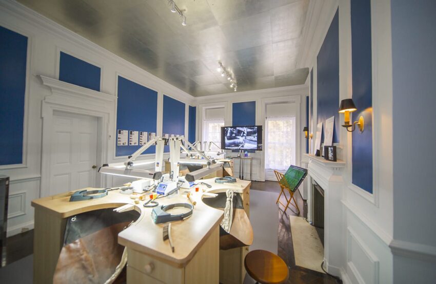 Jewelry workshop with blue walls, workstations, tools, and a monitor displaying designs in a bright room.