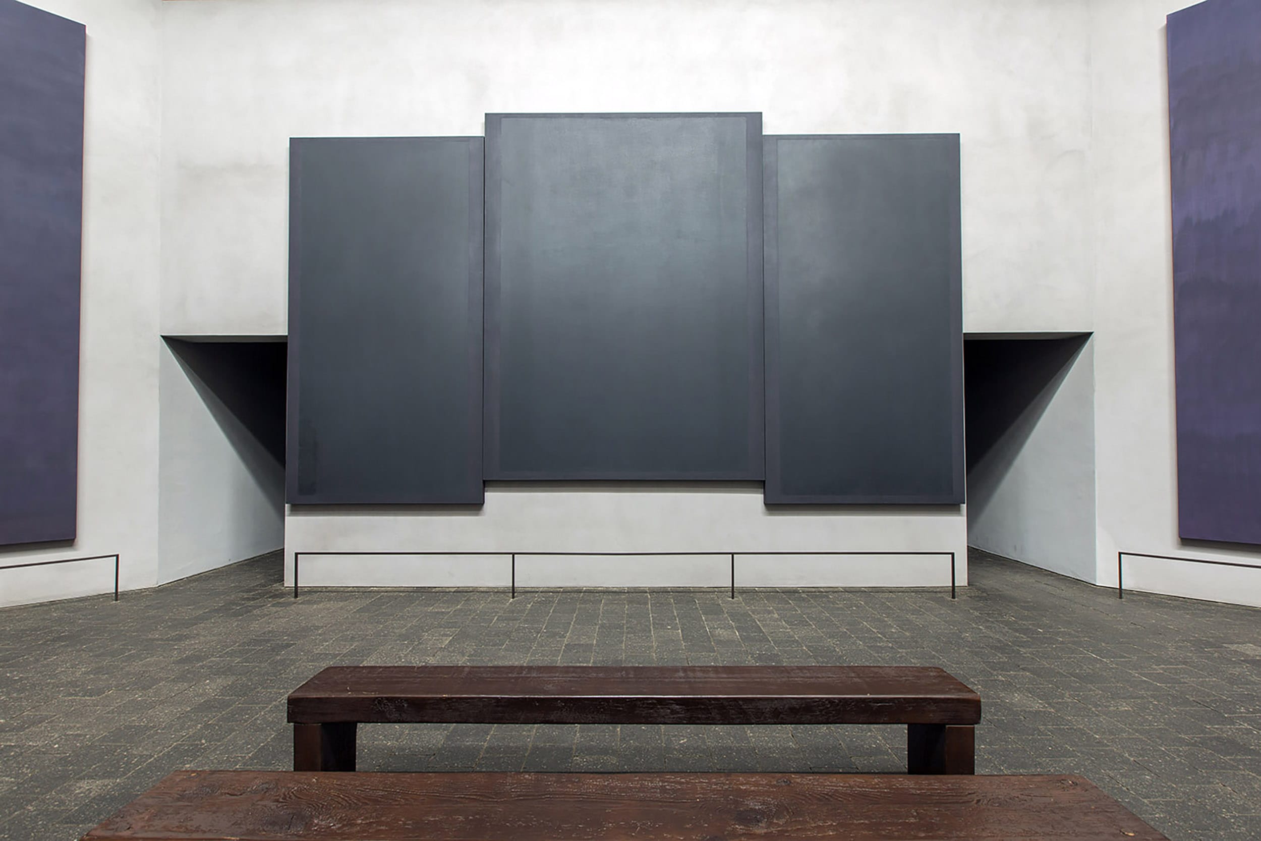 Interior view of a chapel featuring large dark rectangular abstract paintings and a wooden bench in the foreground.
