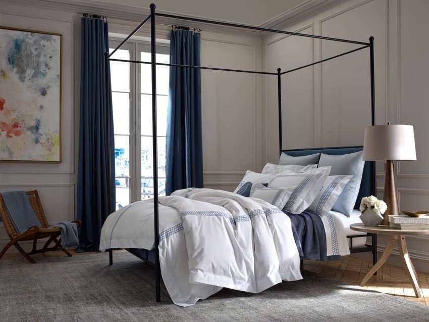 Modern bedroom with a four-poster bed, white bedding, blue accents, a floor lamp, and abstract art on the wall.