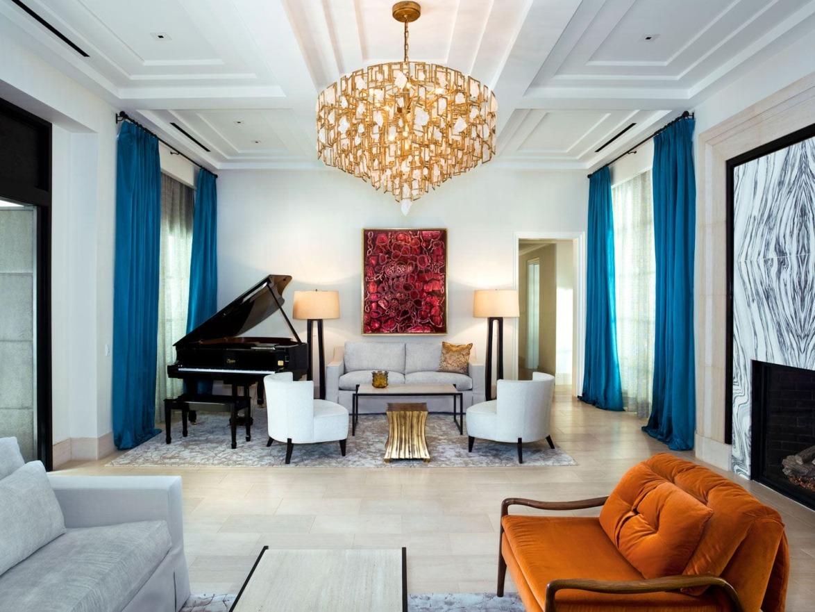 Modern living room with grand piano, blue curtains, orange sofa, chandelier, abstract art, and elegant decor.