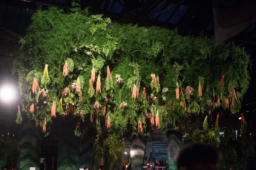 Hanging installation of green foliage with pink-orange flowers under a glass roof.