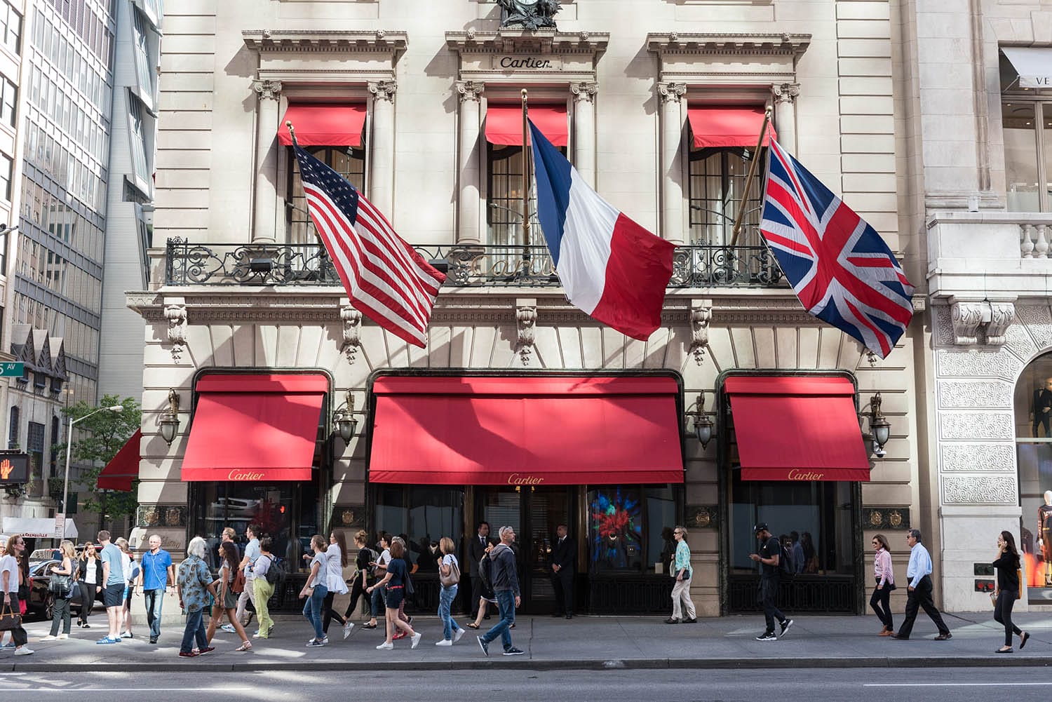 Historic building facade with U.S., French, and U.K. flags; pedestrians walking on the sidewalk in a bustling city area.
