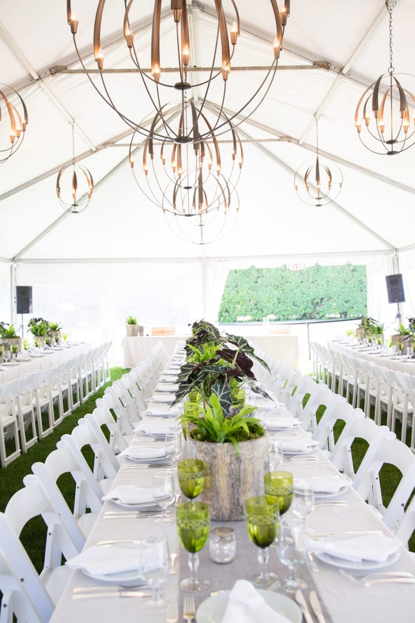 Elegant wedding reception tent setup with long tables, white chairs, green decorations, and ornate chandeliers.