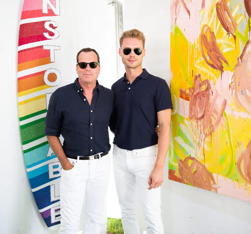 Two men in sunglasses standing next to a colorful surfboard and abstract painting in a sunny outdoor setting.