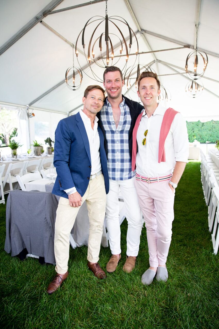 Three men in stylish attire smiling under a tent with white chairs and chandeliers in the background.