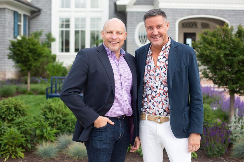 Two men smiling, standing together in a garden with a house in the background.