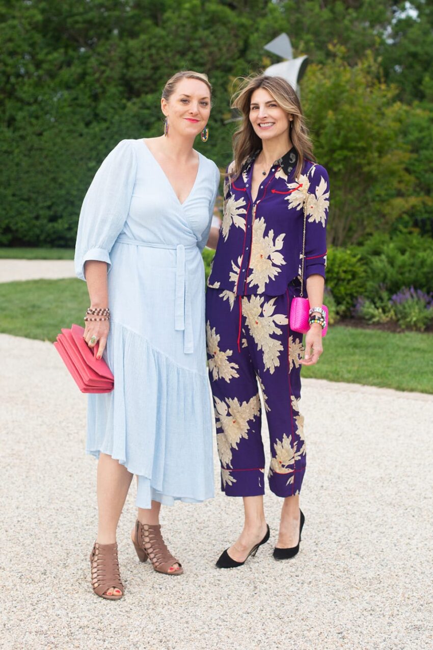 Two women smiling outdoors, one in a light blue dress and the other in a purple floral outfit, holding clutches.