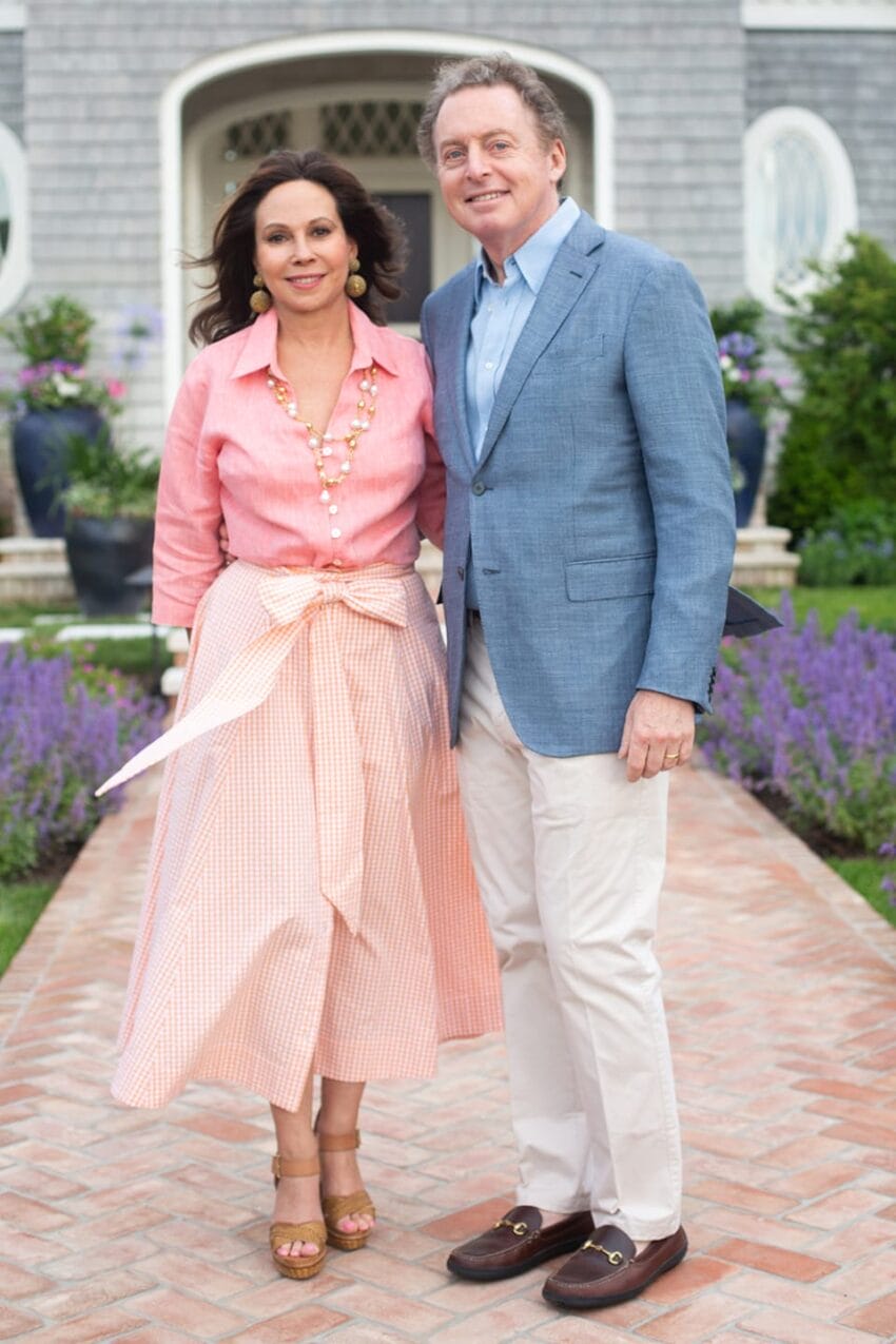 A couple standing on a brick path in front of a house with purple flowers in the background.
