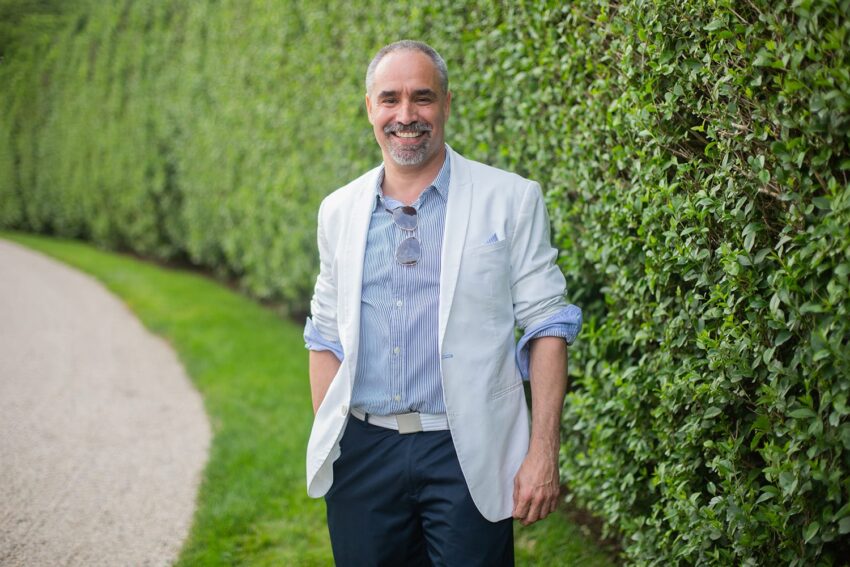 Man in a white blazer smiling while standing on a path next to a tall green hedge.