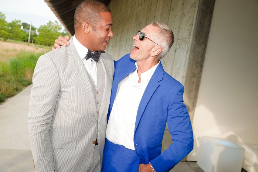Two men in suits smiling at each other outside, one in gray with a bow tie and the other in bright blue with sunglasses.