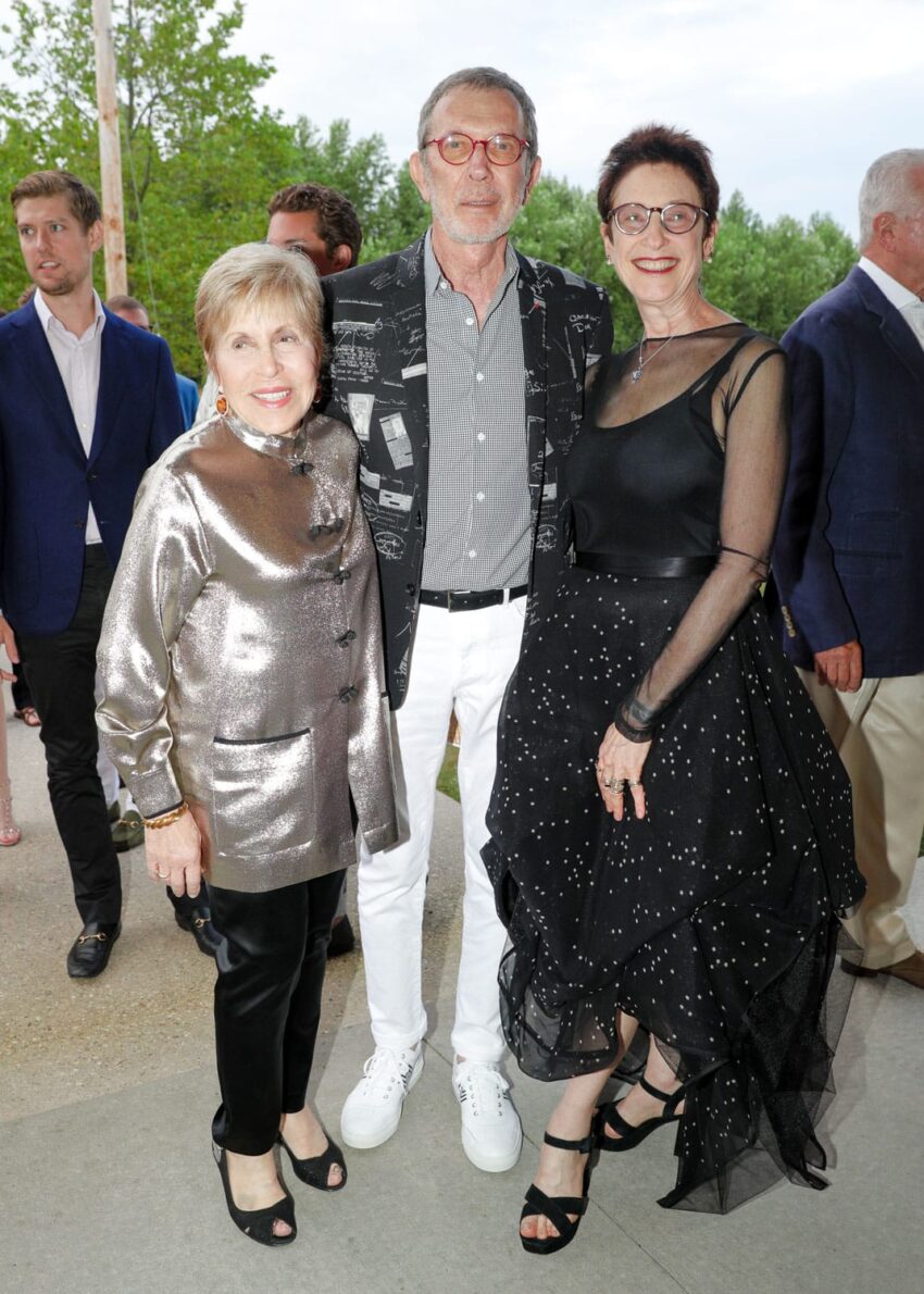 Group of three people smiling and posing outdoors at an event, with trees and other people in the background.