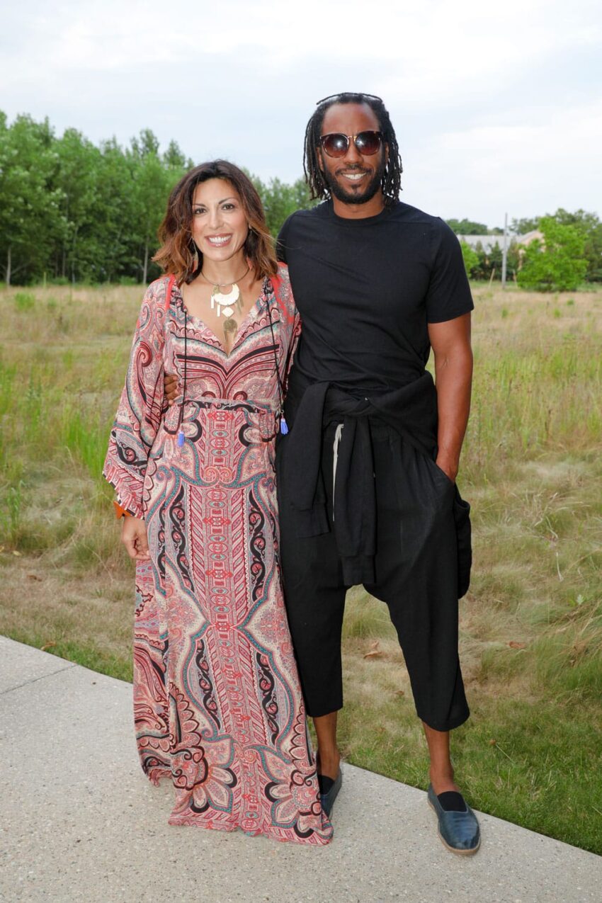 A woman in a patterned dress and a man in black clothing smiling outdoors with greenery in the background.