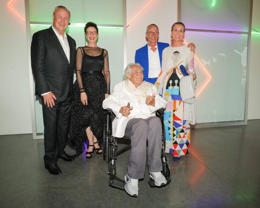 Group of five people posing indoors with colorful lights in the background, one person seated in a wheelchair.
