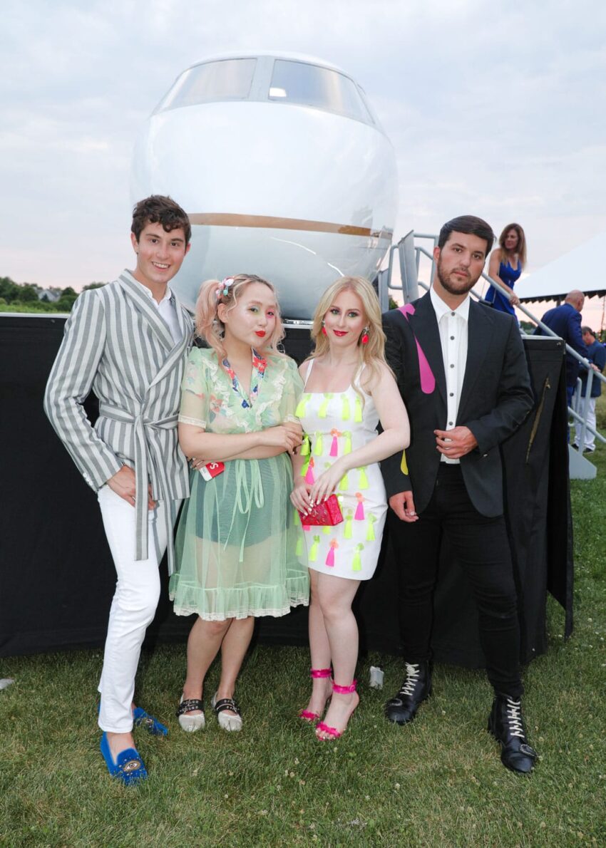 Group of four stylishly dressed individuals posing outdoors in front of a small private jet on a grassy area.