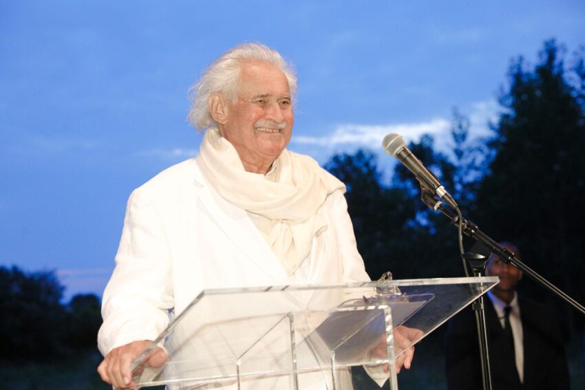 An elderly man in a white suit smiles while standing at a clear podium with a microphone outdoors.