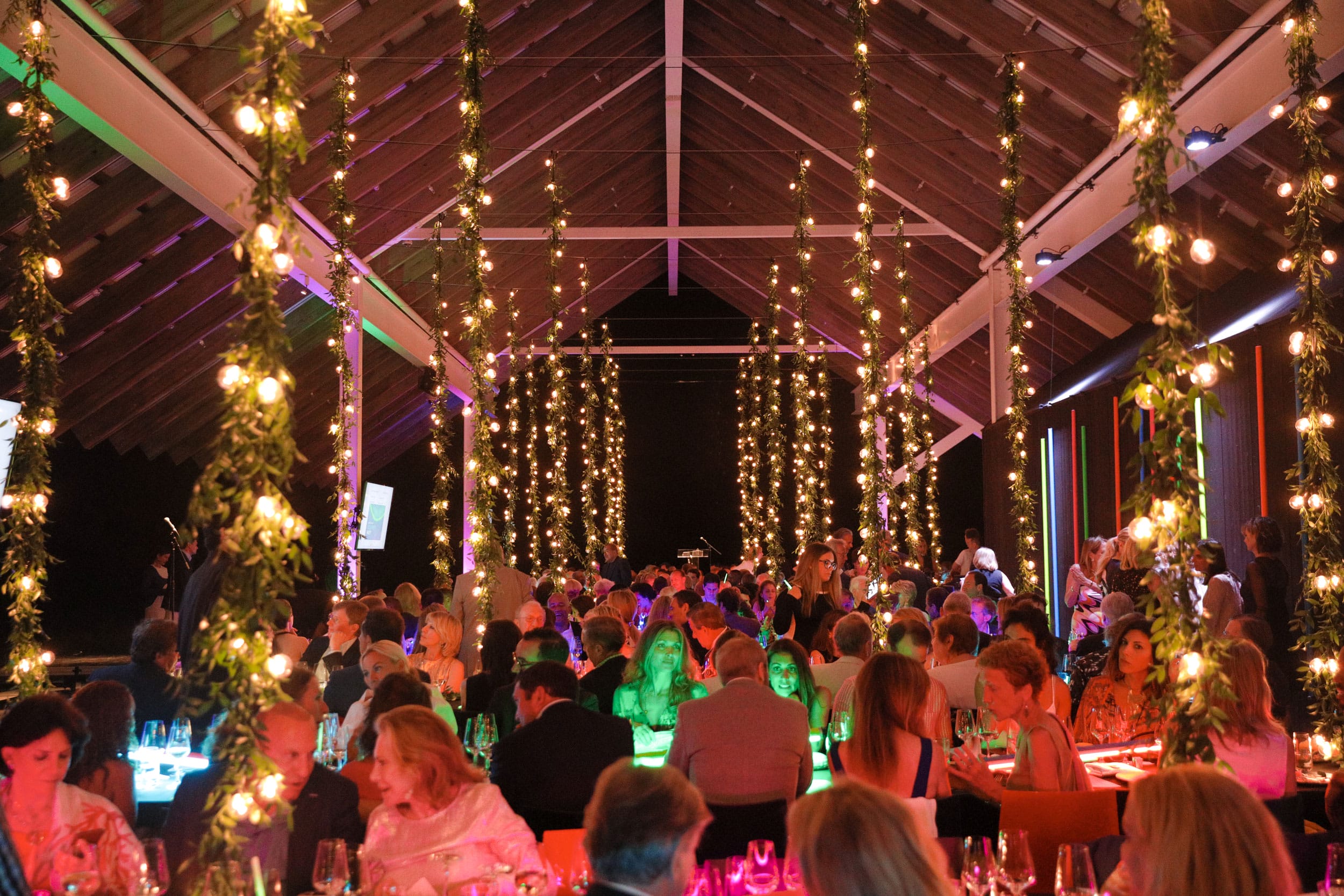 Guests dining at an elegantly lit event under a wooden roof, surrounded by vertical string lights and greenery