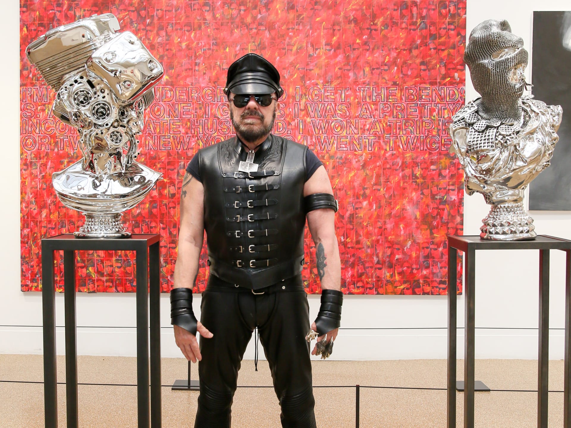 A person in leather attire stands between two metallic sculptures with a vibrant red artwork in the background.