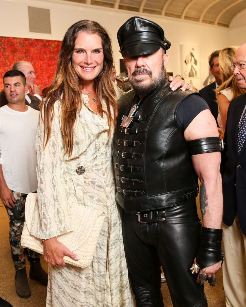 Woman in a patterned dress posing with a man in a leather outfit at an indoor event with people in the background.